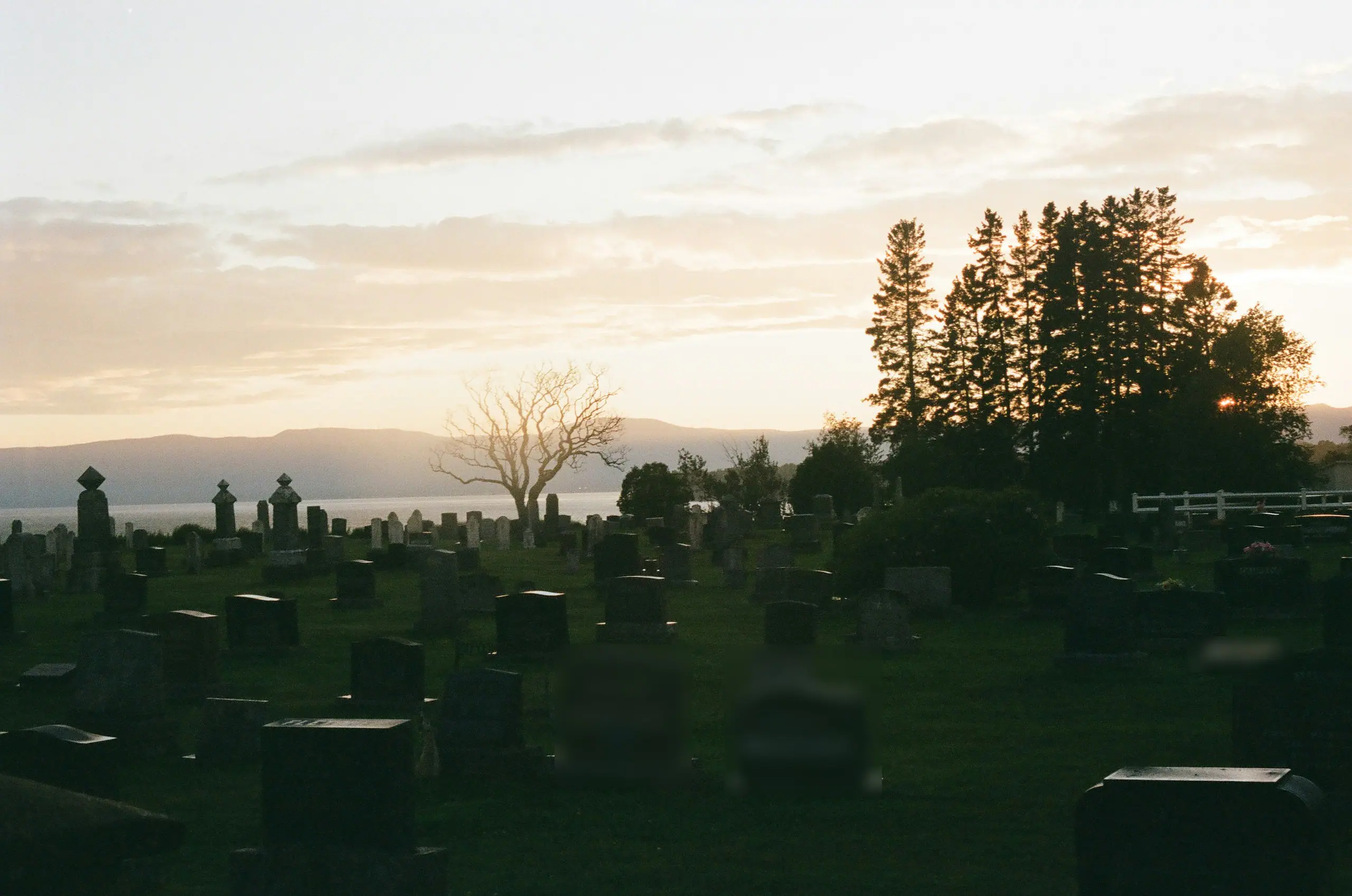 Friedhof über einem See im warmen Abendlicht, dahinter sanfte Hügel